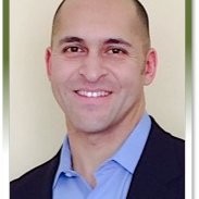 A profile of a man with short dark hair, wearing a blue button up shirt and black jacket.